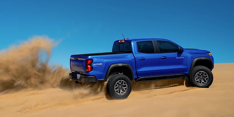 A blue 2025 Chevrolet Colorado driving through a sandy desert, kicking up dust behind it. The truck is shown from the side, highlighting its off-road tires and rugged design against a clear blue sky.