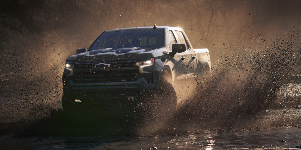 he 2026 Chevrolet Silverado 1500 driving through muddy terrain, kicking up dirt and water, showcasing its off-road capabilities.
